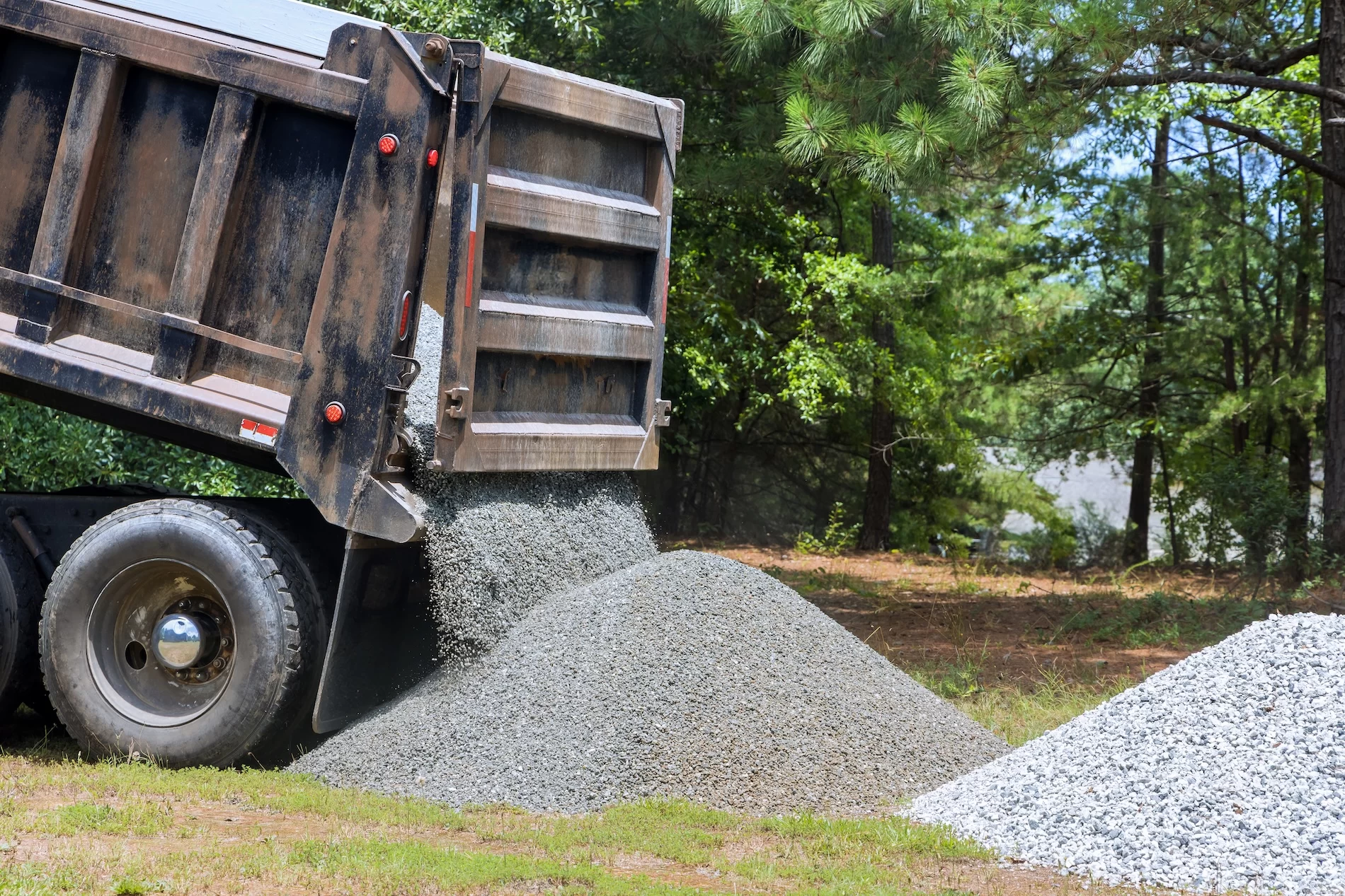 Dump truck actively unloading gravel aggregate material at construction site with forested backdrop